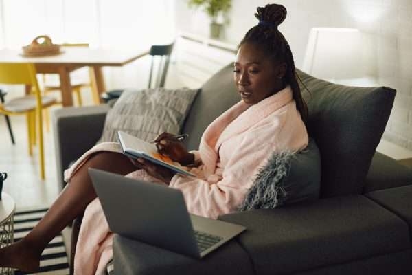 Young black woman in bathrobe writing notes while working on laptop at home.