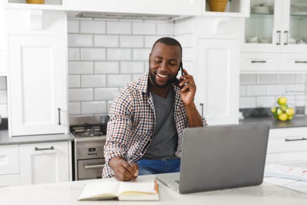 Smiling african american man sitting in kitchen working using laptop and smartphone